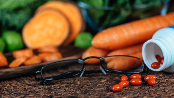 Fekete keretes szemüveg, mellette felborult vitaminos tégelyből kigurult kapszulák, háttérben sárgarépa, zöld leveles zöldségek.