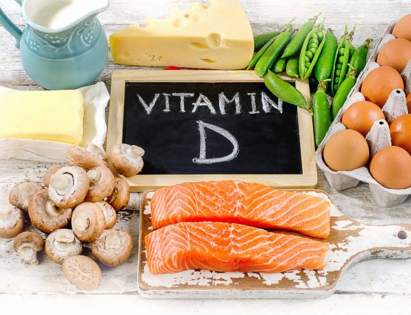 Fehér kopott asztalon egy fekete "Vitamin D" feliratú tábla, körülötte D-vitamin-tartalmú táplálékok: lazac, tojás, sajt, zöldborsó, gomba, tej, vaj.