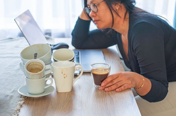 Szemüveges nő könyököl a laptop előtt, a kezénél egy pohár kávé, mellette üres kávéscsészék, bögrék különböző méretben.
