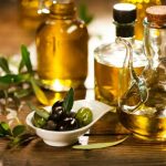 Faasztalon különböző formájú üvegekben olívaolaj, kis fehér tálkában olajbogyók, mellette olajfalevelek.