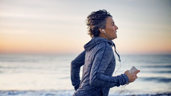 Középkorú nő sportruházatban fut a víz mellett telefonnal a kezében és fülhallgatóval a fülében.