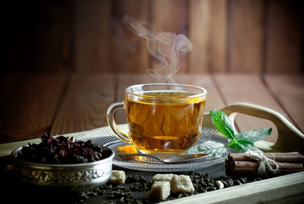 Tálcán üvegcsészében gőzölgő tea, mellette fahéjrudak átkötve, mentalevél, kanál, teafű, barnacukor-darabok.