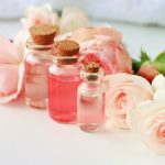 Három különböző méretű üvegcsében rózsavíz, mögöttük rózsaszínű rózsák fekszenek.