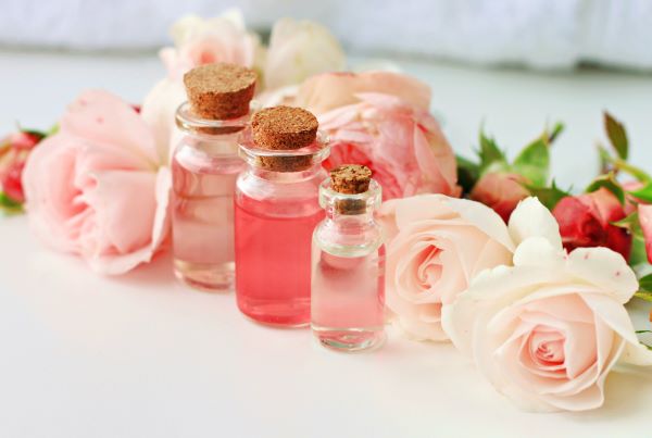 Három különböző méretű üvegcsében rózsavíz, mögöttük rózsaszínű rózsák fekszenek.