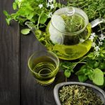 Sötét faasztalon zöld növény, üvegkancsóban zöld tea, pohárban zöld tea, kis tálban szárított teafű.