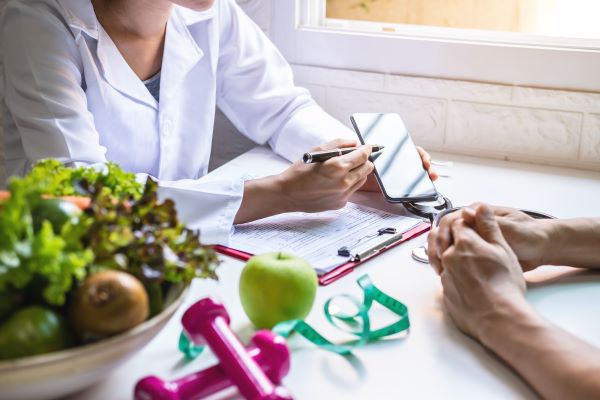 Asztalnál ülő fehér köpenyes doktornő magyaráz a szemben ülő páciensnek, az asztalon mappa, egy tál zöldség, alma, kézi súlyzó, mérőszalag.