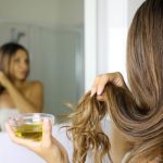 Hosszú hajú nő áll a fürdőszobatükör előtt, egyik kezével egy hajtincsét fogja, másikban kis üvegtálka olajat tart.