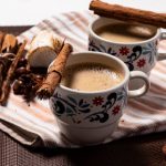 Csíkos konyharuhán két virágmintás bögrében kávé, tetejükön egy-egy rúd fahéj, mellette egy köteg fahéjrúd, egy kanál kakaópor.