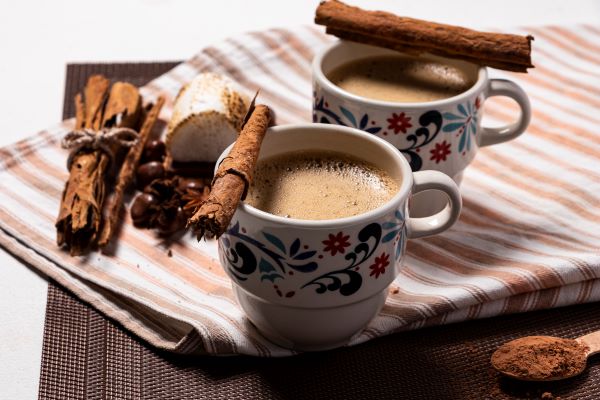 Csíkos konyharuhán két virágmintás bögrében kávé, tetejükön egy-egy rúd fahéj, mellette egy köteg fahéjrúd, egy kanál kakaópor.