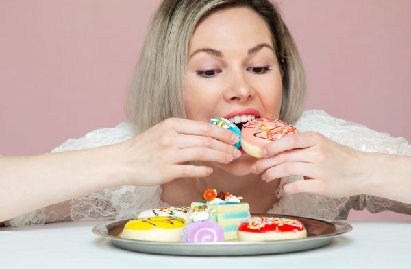 Fiatal nő színes süteményekkel teli tányér előtt ülve egyszerre két süteményt próbál a szájába tenni.