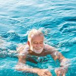 Kék vizű úszómedencében idős, szakállas férfi úszik mosolyogva.