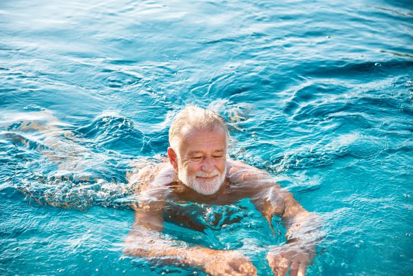 Kék vizű úszómedencében idős, szakállas férfi úszik mosolyogva.