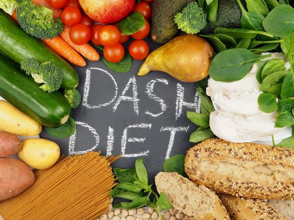 Fekete alapon fehér "DASH-diéta" felirat, körülötte zöldségek, gyümölcsök, teljes kiőrlésű tészta és péksütemények.