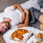 Fiatal, szakállas férfi fekszik az ágyon hasát fogva, előtte pizza, hamburger, sült krumpli.