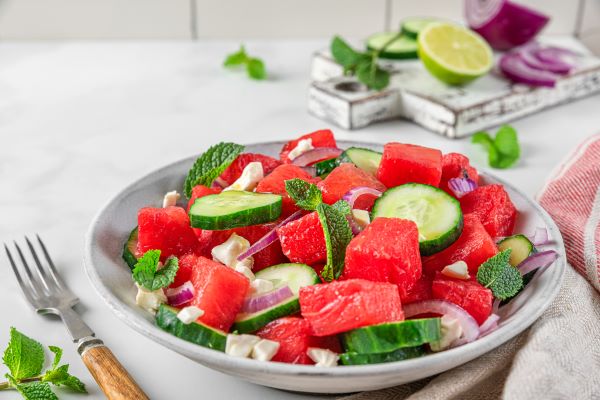 Tányéron görögdinnye-saláta uborkával, lila hagymával, háttérben fehér koptatott vágódeszkán lime és lila hagyma.