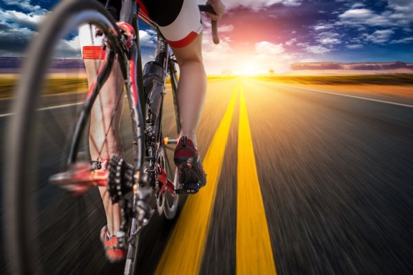 Biciklin ülő nő lábai, előtte az út, amelynek végén kel fel a nap.