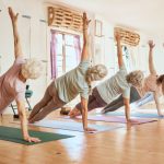 Idős nők jógáznak egy teremben tornaszőnyegeken.