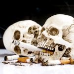 Egy emberi koponya szájában égő cigaretta, mellette elnyomott cigarettacsikkek és egy eldőlt koponya.
