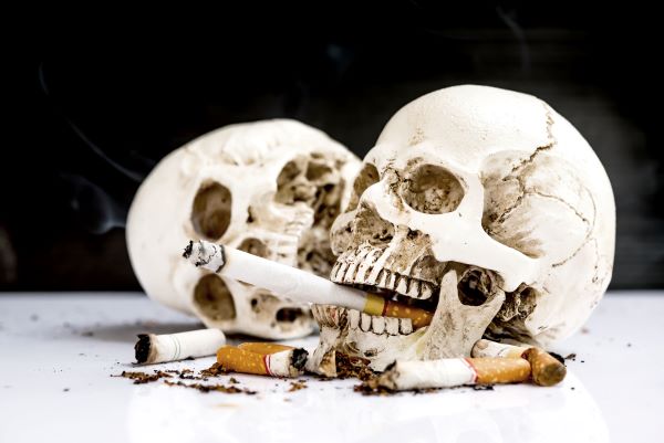 Egy emberi koponya szájában égő cigaretta, mellette elnyomott cigarettacsikkek és egy eldőlt koponya.