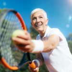 Idősebb nő teniszezik, háttérben fák, kék ég.