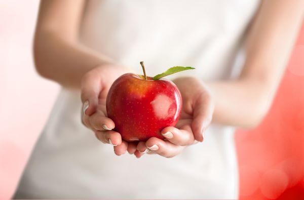 Fehér ruhás nő két kézzel tart egy piros almát.
