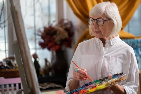 Idős nő festékpalettával a kezében áll a festővászon előtt.