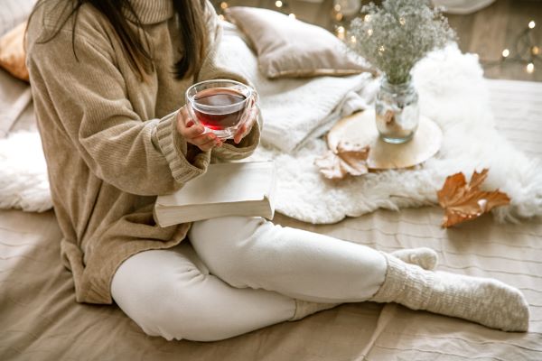 Nő pulóverben, vastag zokniban, párnákkal és szőrmével körülvéve ül egy könyvvel az ölében és egy csésze teával a kezében.