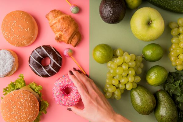 Baloldalt rózsaszínű alapon egészségtelen ételek, jobboldalt zöld alapon zöld gyümölcsök, egy női kéz nyúl egy fánkért.