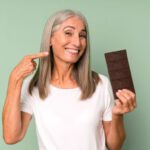 Hosszú ősz hajú idős nő széles mosollyal mutat a másik kezében lévő tábla csokoládéra.