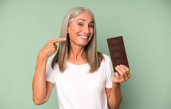 Hosszú ősz hajú idős nő széles mosollyal mutat a másik kezében lévő tábla csokoládéra.