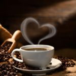 Fehér csészében gőzölgő kávé szív alakú gőzzel, mellette zsák kávébabbal és mérőkanállal.
