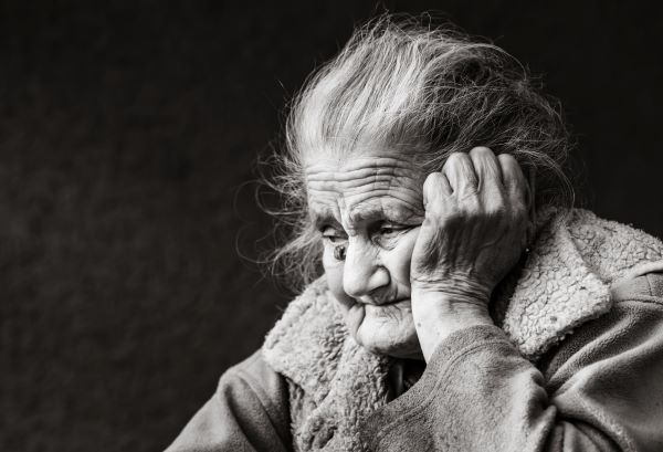 Fekete-fehér fotón nagyon öreg néni kabátban könyököl és szomorúan néz maga elé.