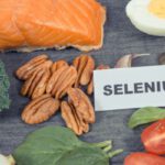 Szeléntartalmú táplálékok "selenium" feliratú táblával: lazac, főtt tojás, paradicsom, brazil dió, fokhagyma, tökmag, brokkoli, spenót.