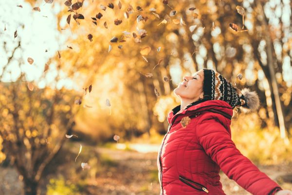 Fiatal nő sapkában és piros kabátban élvezi az őszi természetet, miközben hullanak rá a falevelek.