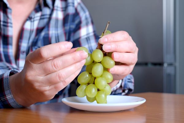 Asztalnál ülve kis fehér tányérból valaki kockás ingben szőlőt eszik.