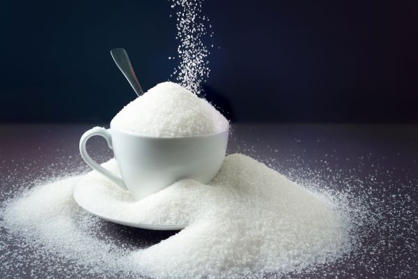 Fehér csésze megpúpozva kristálycukorral, közben valaki szórja még rá a cukrot.
