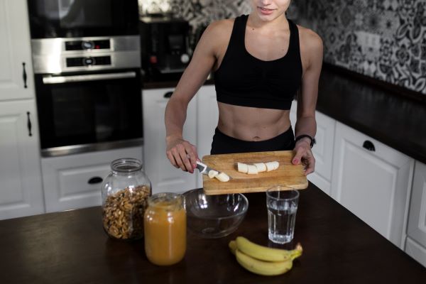 Fiatal nő edzőruhában a konyhában szeletelt banánt tesz egy üvegtálba, mellette a pulton banán, pohár, nagy befőttesüvegben dió.