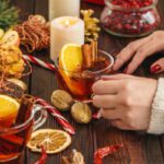 Faasztalon női kezek üvegcsészét fognak, benne narancsos fahéjas teával, mellette gyertya, aranyszínű dió, cukorka, szárított narancsszeletek.