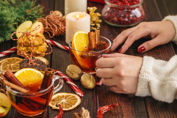 Faasztalon női kezek üvegcsészét fognak, benne narancsos fahéjas teával, mellette gyertya, aranyszínű dió, cukorka, szárított narancsszeletek.