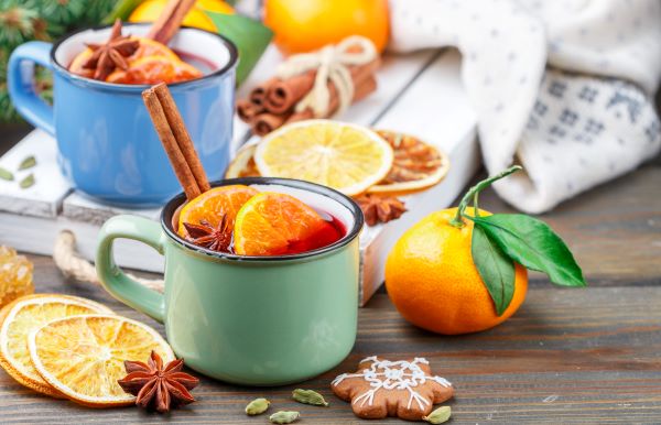 Színes bögrékben tea mandarinszeletekkel, fahéjrúddal, körülötte szárított narancsszeletek, csillagánizs, kardamom, mézeskalács, egy köteg fahéjrúd.