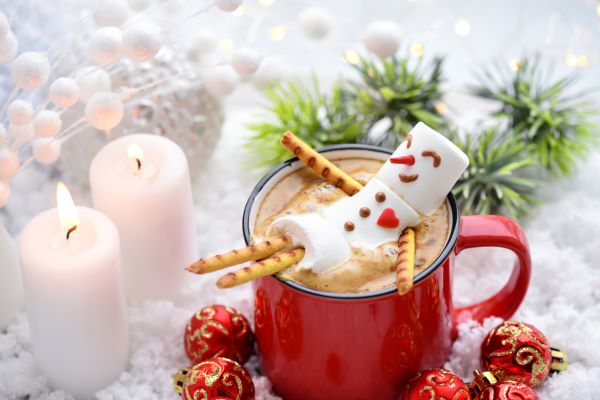 Piros bögrében kávé hóember-pillecukorral, mellette gyertyák, fenyőág, karácsonyfagömbök.
