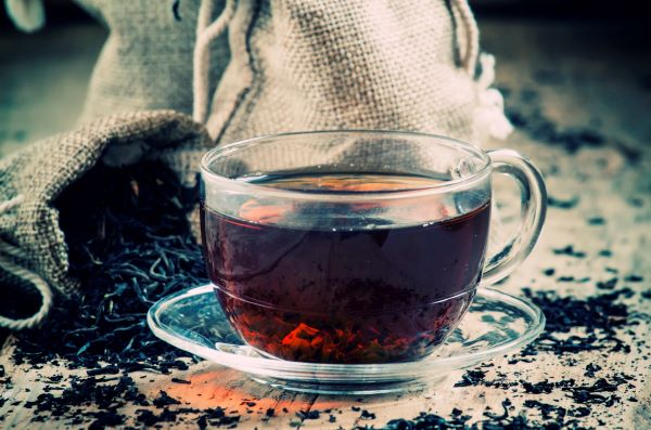 Üvegcsészében fekete tea, körülötte szárított tealevelek szétszórva, illetve kis vászonzsákokban feketetea-levél.