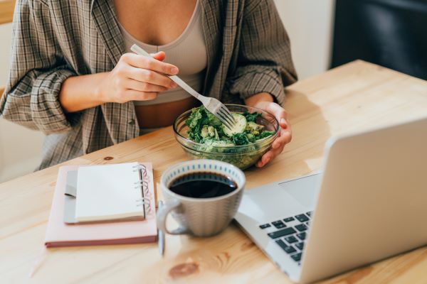 Nő kockás ingben ül a laptop előtt, miközben üvegtálból salátát eszik, előtte egy csésze kávé, jegyzetfüzet.
