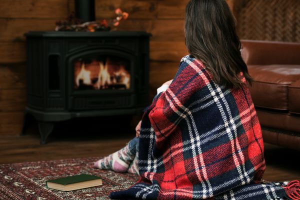 Barna hosszú hajú nő kockás plédbe burkolózva ül a szőnyegen a kályha előtt, amelyben lobog a tűz.