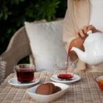 Nő fehér teáskannából üvegcsészékbe teát tölt kerti fonott asztalnál ülve, mellette egy másik nő ül.