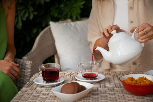 Nő fehér teáskannából üvegcsészékbe teát tölt kerti fonott asztalnál ülve, mellette egy másik nő ül.