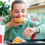 Fiatal nő gyorsétteremben óráját nézve eszik hamburgert, előtte az asztalon sült krumpli és üdítő.