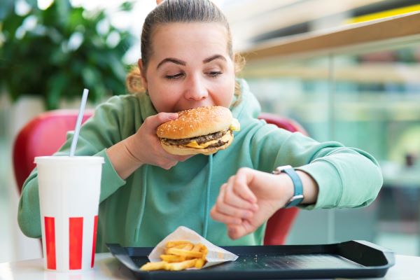 Fiatal nő gyorsétteremben óráját nézve eszik hamburgert, előtte az asztalon sült krumpli és üdítő.