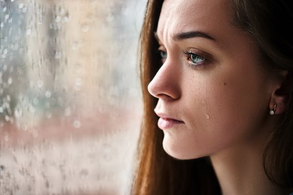 Fiatal nő könnyes szemmel áll a vizes ablaküvegnél.