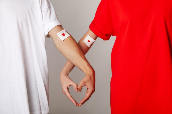 Egy fehér pólós férfi és piros pólós nő karja könyökhajlatban leragasztva, a gézen kis piros szívvel, karjaik összefonódva szívet formálnak.